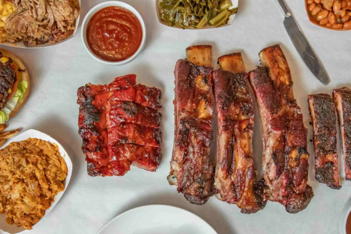 A spread of Redbones barbecue on white paper: a slab of pork ribs, three long beef ribs, pulled pork, chopped burnt ends, a cheeseburger, collard greens, baked beans and a bowl of sauce
